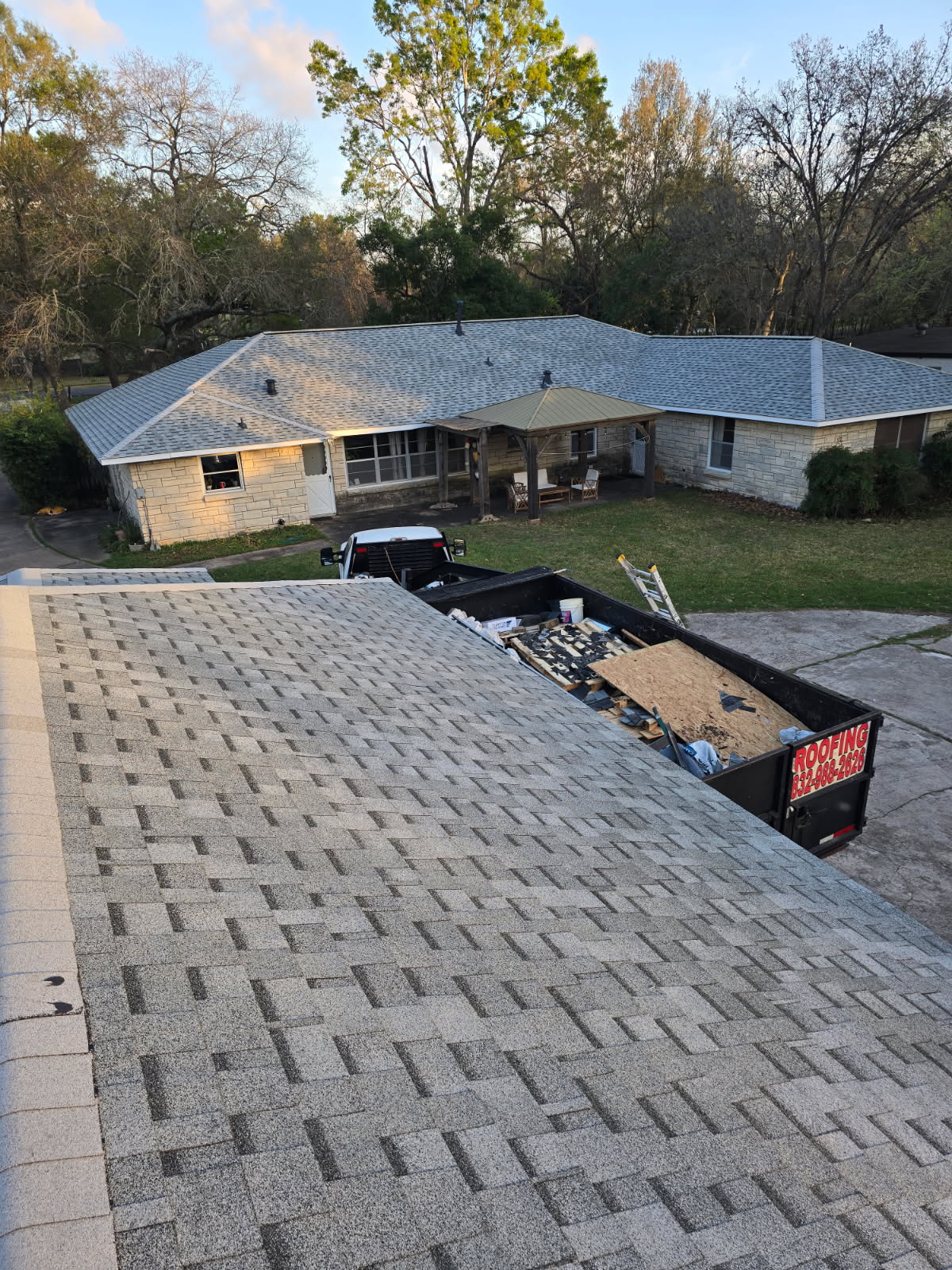 View across a shingled roof slope toward a house with a light grey shingle roof and a roofing dumpster.