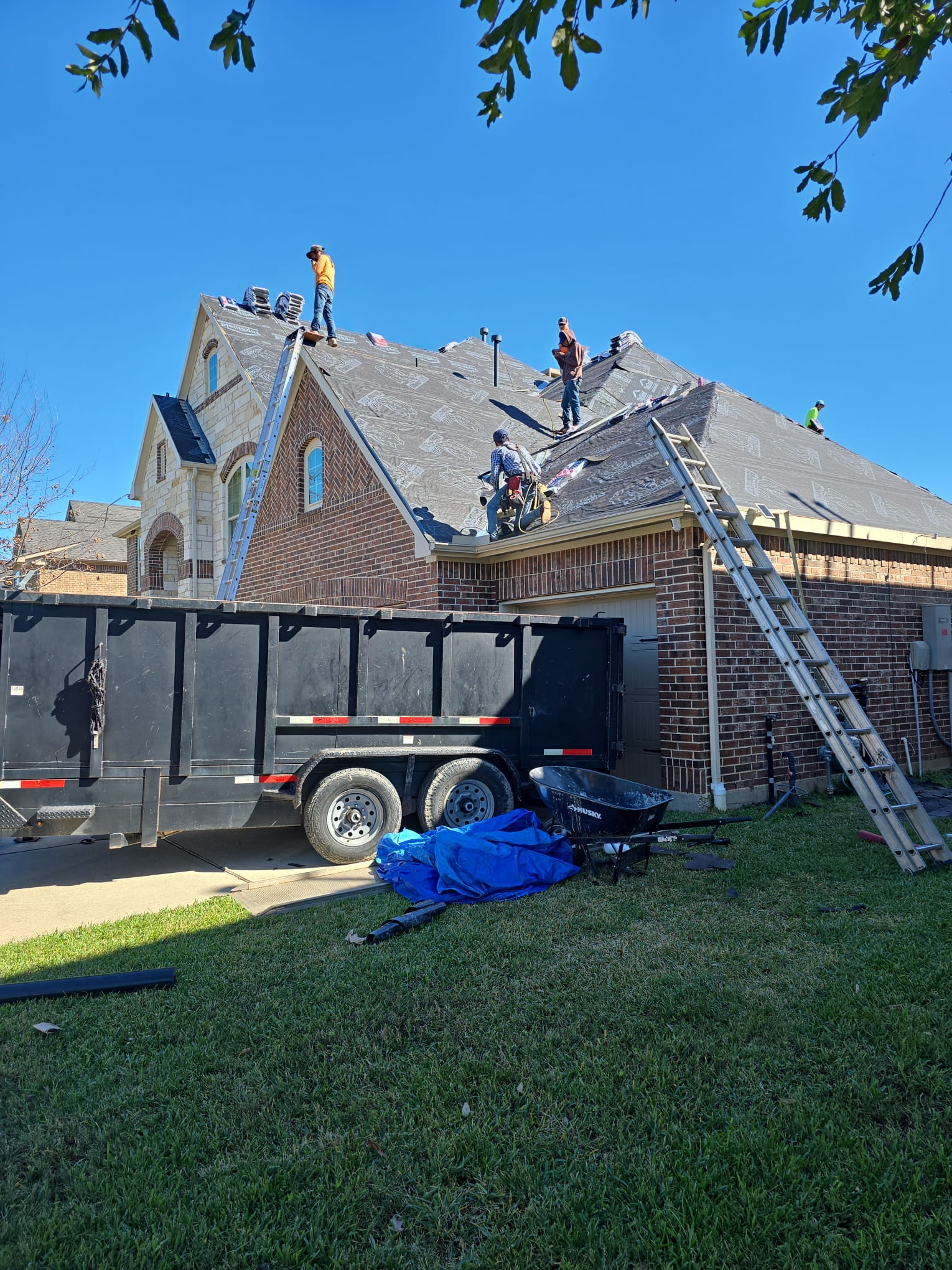 Crew on a roof covered in underlayment, with ladders and a loaded debris trailer in the driveway.