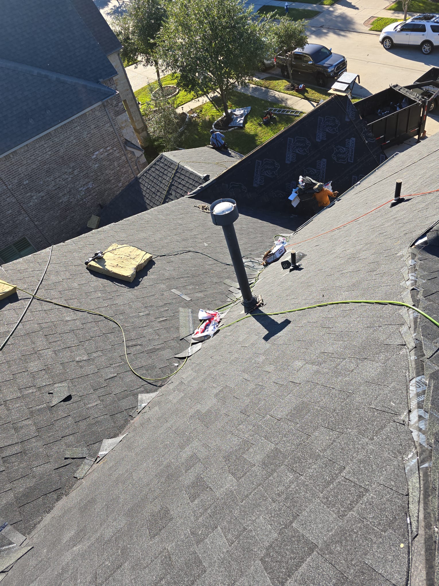 Elevated view over shingle roof slopes with a tall vent pipe and a dumpster in the street below.