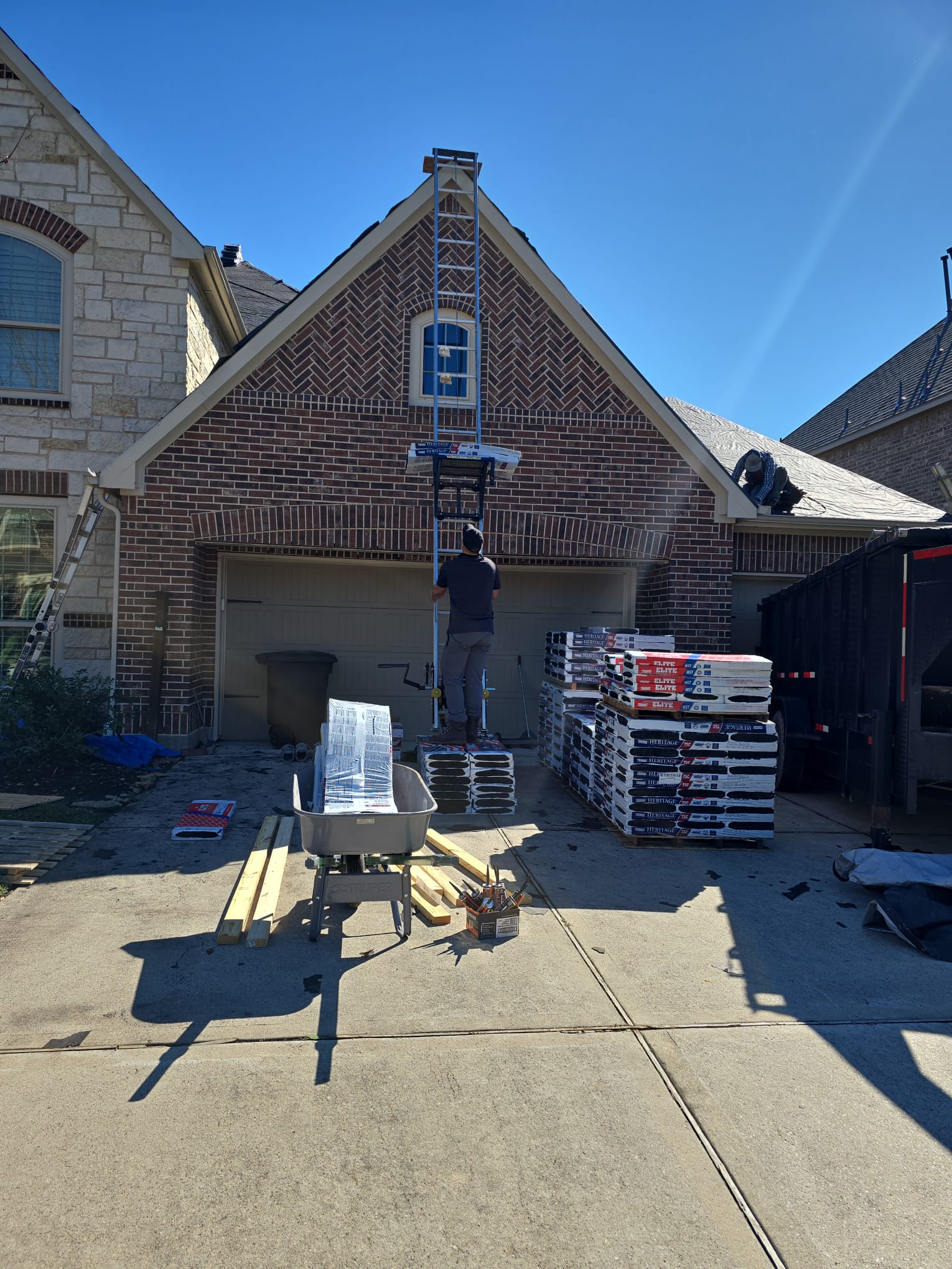 Worker on a ladder hoist lifting shingle bundles to the roof, with pallets of shingles in the driveway.