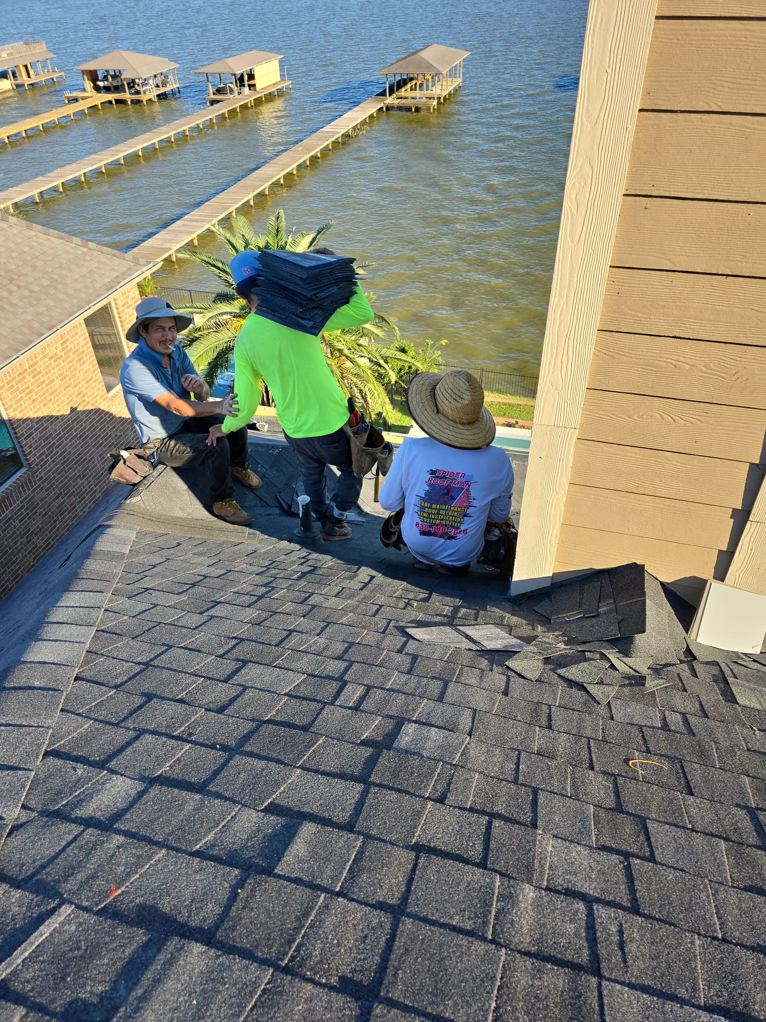 Three roofers on a shingle roof beside a siding wall, one carrying a bundle of shingles.