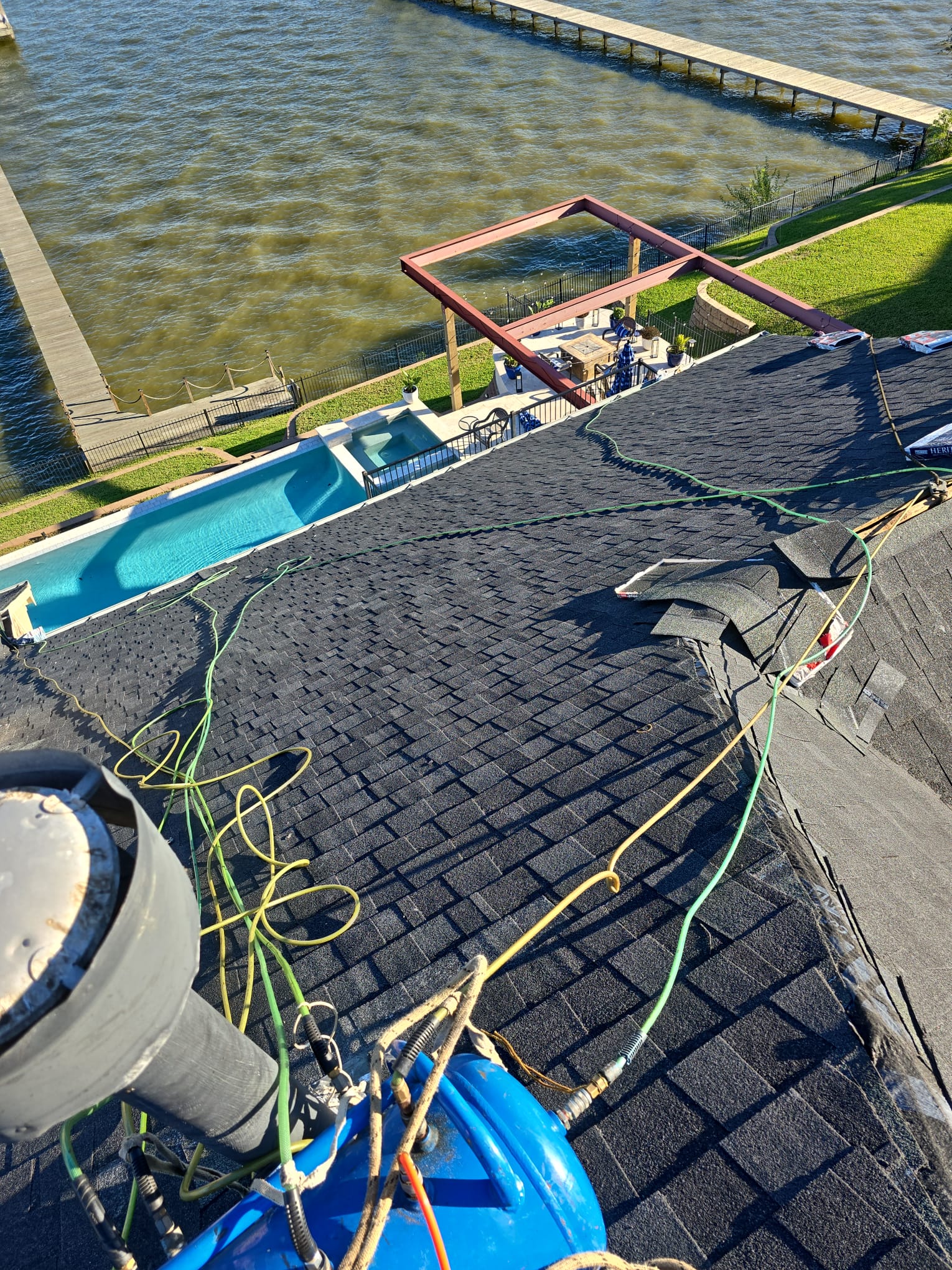 Shingle roof slope with hoses and materials, overlooking a pool and a waterfront dock.