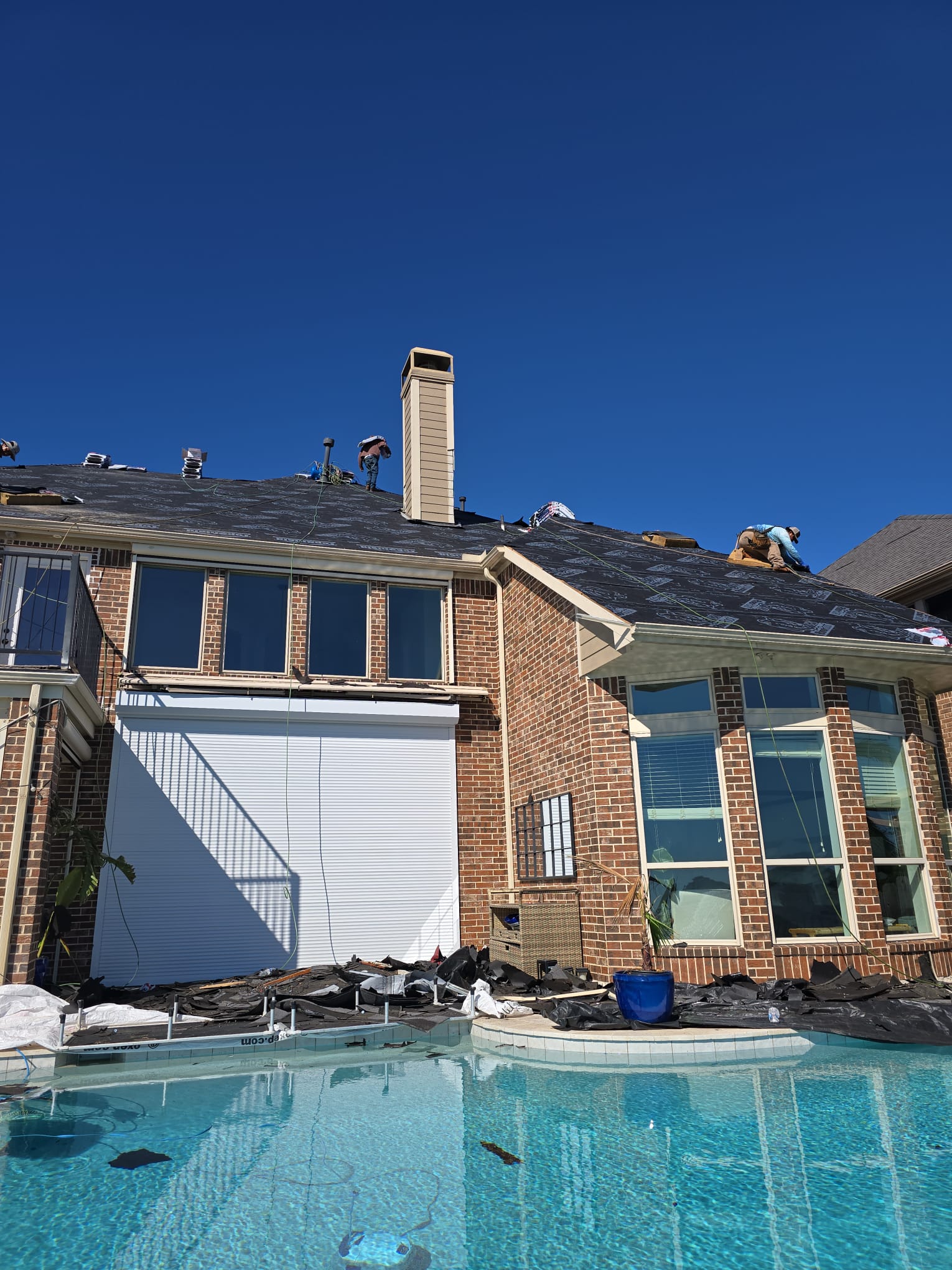 Two-storey brick house with roofers working on the underlayment-covered roof, seen from the pool.