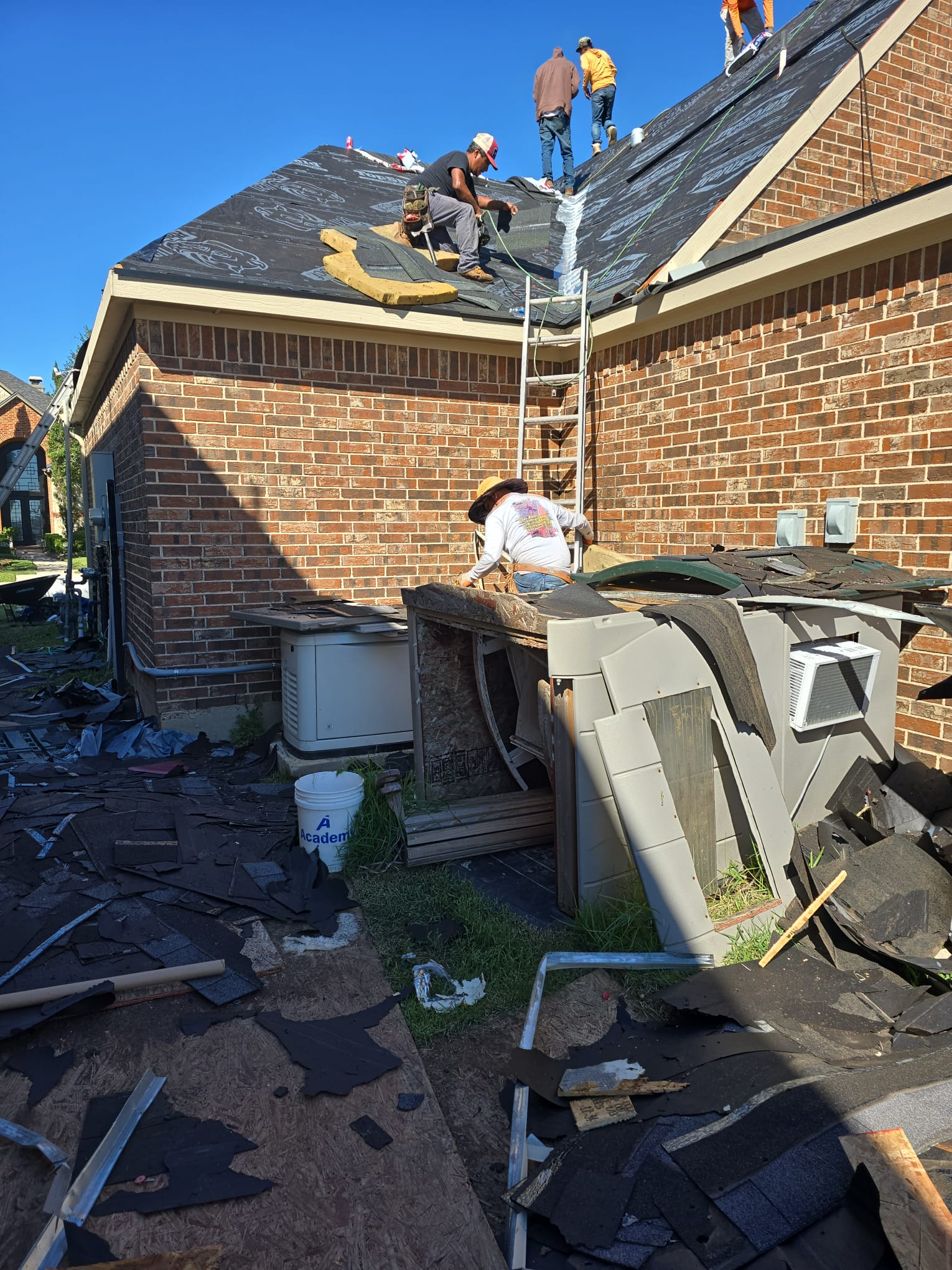 Crew tearing off the old covering beside a brick house, with old shingles cleared onto the ground.